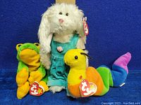 Three Ty Beanie Babies plush toys: white rabbit in teal overalls with pink flower, green and yellow frog, and multi-colored worm named Inch