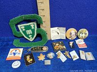 Full view of the lot showing a green varsity sweater letter patch, various pin back buttons and badges including Halloween and animal themes, small enamel pins and metal badges, all arranged on blue background next to a ruler.