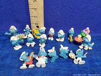 Photo of 15 vintage Smurf figurines from 1970s-80s, showing various characters in different poses with accessories, placed in front of a ruler for size reference.
