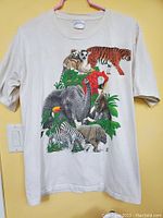 Front view of cream-colored short sleeve t-shirt with colorful African animal illustration including lemurs, tiger, elephant, zebra, rhinoceros, bird, koala, and foliage.