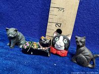 Four miniature metal cat figurines displayed side by side with a ruler for size reference. Includes one black with floral pattern, one black and white with red bow tie, and two gray textured cats.