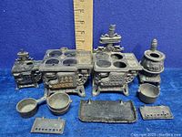 Four cast iron miniature Queen stoves of different sizes, with accompanying pots and trivets, displayed against a blue background with visible ruler for scale.