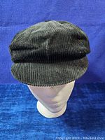 Front view of a black corduroy newsboy cap on a white mannequin head against a blue background, showing the front brim and textured fabric.