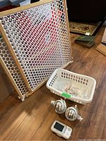 Photo showing white child safety gate with honeycomb plastic center and wooden frame, plastic basket with power cords, and two silver child safety cameras with mounts and one parent monitor screen.