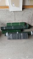 Several green plastic barrier fencing rolls and a roll of metal wire mesh placed on concrete floor against a wall.