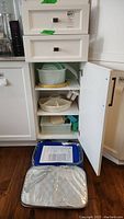 Arctic Zone insulated silver food carrier bag shown open on floor in front of white cabinet with shelves containing white ceramic and plastic serving dishes and various containers.