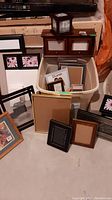 Wide view of assorted wooden and metal picture frames with some framed photos and blank mats, placed around a plastic bin