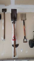 Photo of three hanging garden tools: two shovels and a pitchfork with wooden handles against a wall.