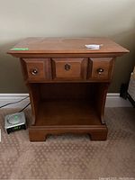 Front view of one Vilas brand wooden night stand with single drawer and lower open shelf, showing round metal knobs and keyhole detail. Positioned on carpet with adjacent electrical items.