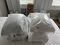 Four bed pillows: two in striped pillowcases and two wrapped in clear plastic covers, positioned near a window.