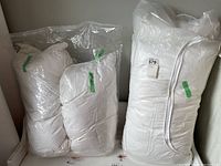 Four king size pillows; two in white pillowcases laid out on a bed, one showing slight creases and implied use, two still wrapped in plastic packaging standing upright against a wall.