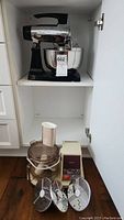 Sunbeam black stand mixer in cabinet with stainless steel mixing bowl and Philips food processor motor and attachments on floor.