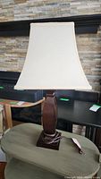 Full view of the wooden base table lamp with white fabric shade on a greenish surface in a room with stone tile wall.