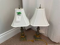 Pair of table lamps shown side by side, exhibiting white pleated shades and antique-style bases with yellow cords coiled around them.
