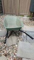 Green fiberglass bucket wheelbarrow with black metal handles and frame placed on a rock and stone surface outside near a wooden fence.