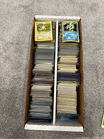 A long cardboard box filled with thousands of Pokémon trading cards divided into two sections with various card faces visible on top.