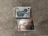 Two NBA trading cards in plastic sleeves on gray carpet, one with teal uniform player and one with white and red uniform player.