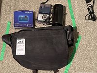 Photo showing the black padded camera bag, boxed Olympus Stylus Tough 3000 digital camera, battery charger, film canister, and cables arranged on carpet.