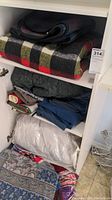 Shelving holding various folded blankets, linens, blue linens, and large white duvet in plastic.