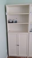 Front view of the white bookcase showing three shelves above two closed cabinet doors with metal knobs. Items placed on middle shelves are excluded from the lot.