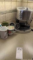 Photo showing the silver DeLonghi coffee maker placed on a kitchen counter with a drip tray and control buttons visible. Five assorted coffee mugs are arranged beside it including white, floral, green, gray, and colorful design mugs.