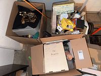 Overhead shot of three open cardboard boxes containing various craft supplies such as brushes, tins, glue bottles, and decorative cords.