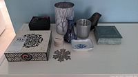 Displays various decorative boxes, candle holders including frosted design, ceramic bird figurine, vases, a small dish, and a snowflake ornament arranged on a table.