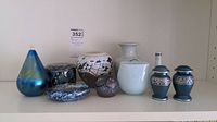Front view of all seven ceramic and glass decorative items arranged on a shelf including vases, bowls, and urns in various colors and finishes.