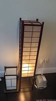 Set of one tall floor lamp and two smaller desk lamps made with wooden frames and translucent white panels, all illuminated, showing size contrast
