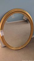 Gold oval framed mirror resting against a wall with carpeted floor, showing full front view.