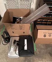 Overview photo showing flatbed scanner with cords, black multifunction printer/scanner, and a cardboard box containing office supplies such as rolled plastic sheets and file folders.