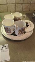 Set of four royal commemorative mugs and a plate displayed on countertop, showing detailed royal crests and images.