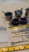 Vintage electroplated silver condiment pots, mustard bowls with blue glass inserts and matching spoons, pepper shaker, and shell-shaped spoon with mother of pearl and silver utensils on a cloth surface