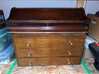 Large roll-top wooden storage box on top of smaller three-drawer wooden chest in a storage room.