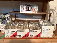 Photo showing three trays of GEMYT wine glasses, each tray containing six clear glass wine glasses, and a boxed gigantic wine glass from BigMouth Inc. on a wooden shelf.