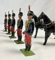 Five standing soldiers in red pants and blue jackets with gold trim, two black horses behind