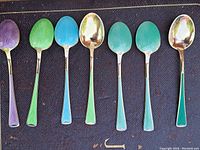 Seven enamelled teaspoons laid out on textured surface in order of color
