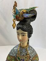 Close-up of figure head showing carved bone face, black hair, elaborate enamel headdress with bird motif and dangling pendant
