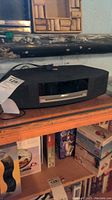Full view of black Bose Wave Music System III speaker sitting on a wooden shelf with power cord attached, showing front panel with display.