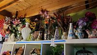 Wide view of artisan ceramics, glass vases, figurines, and faux flowers arranged on a white shelving unit beneath a wooden ceiling with visible beams.