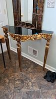 Full view of the ornate gold toned demi lune table with a black mirrored top showing the curved front and detailed legs.