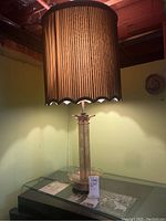 Full view of table lamp on glass surface with tall ridged brown lampshade and base with textured metal and glass bowl section.