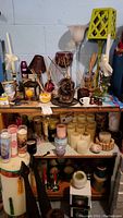 Overview photo of the entire shelf showing multiple candles, candle holders, figurines, reed diffusers, and small decorative lamps in a basement setting.
