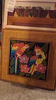 Framed colorful bird painting with natural woven wood frame, surrounded by other framed artworks visible behind