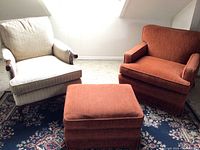 Two fabric armchairs and an ottoman on a patterned rug. One armchair is beige with wooden armrests, the other burnt orange. Ottoman matches orange chair.