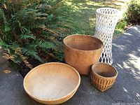 Four baskets displayed outdoors on pavement with greenery in background, showing the entire set including the tall white basket stand.