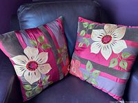 Two square throw pillows with large cream flowers and green leaves on pink, purple, and brown stripe background, placed on a dark chair