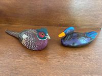 Two hand painted carved bird figurines including a quail and a duck seen from a front angle on wood surface.