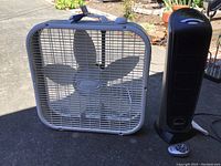 Front view of white Lasko box fan with five blades and black Lasko tower heater with remote on the ground.