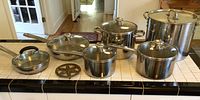 Front view of entire lot showing six stainless steel cookware pieces with glass lids and one vintage metal trivet arranged on tile countertop